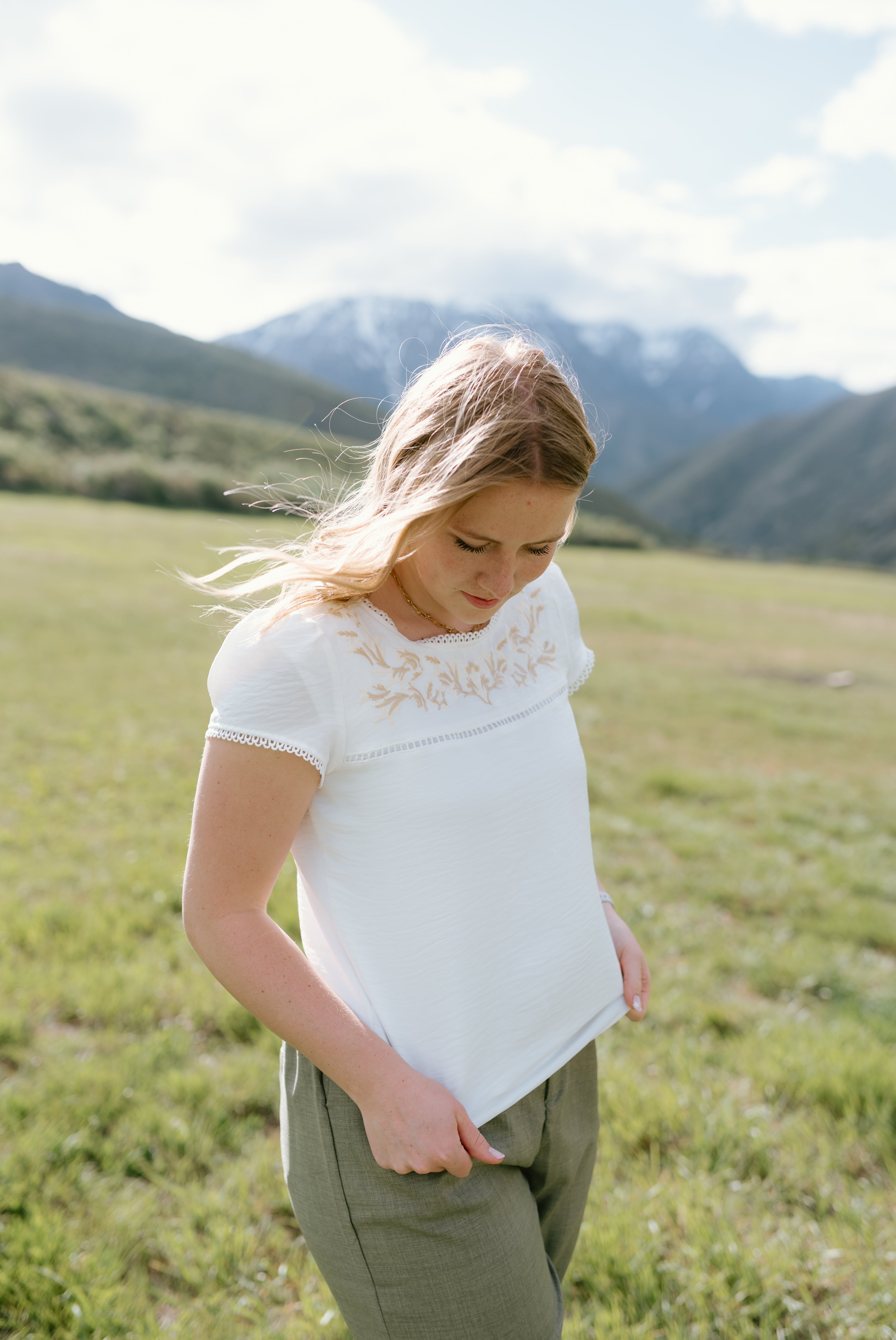 Young woman wearing a white Hampton Top with embroidery, outdoors in a grassy field with mountains in the background.