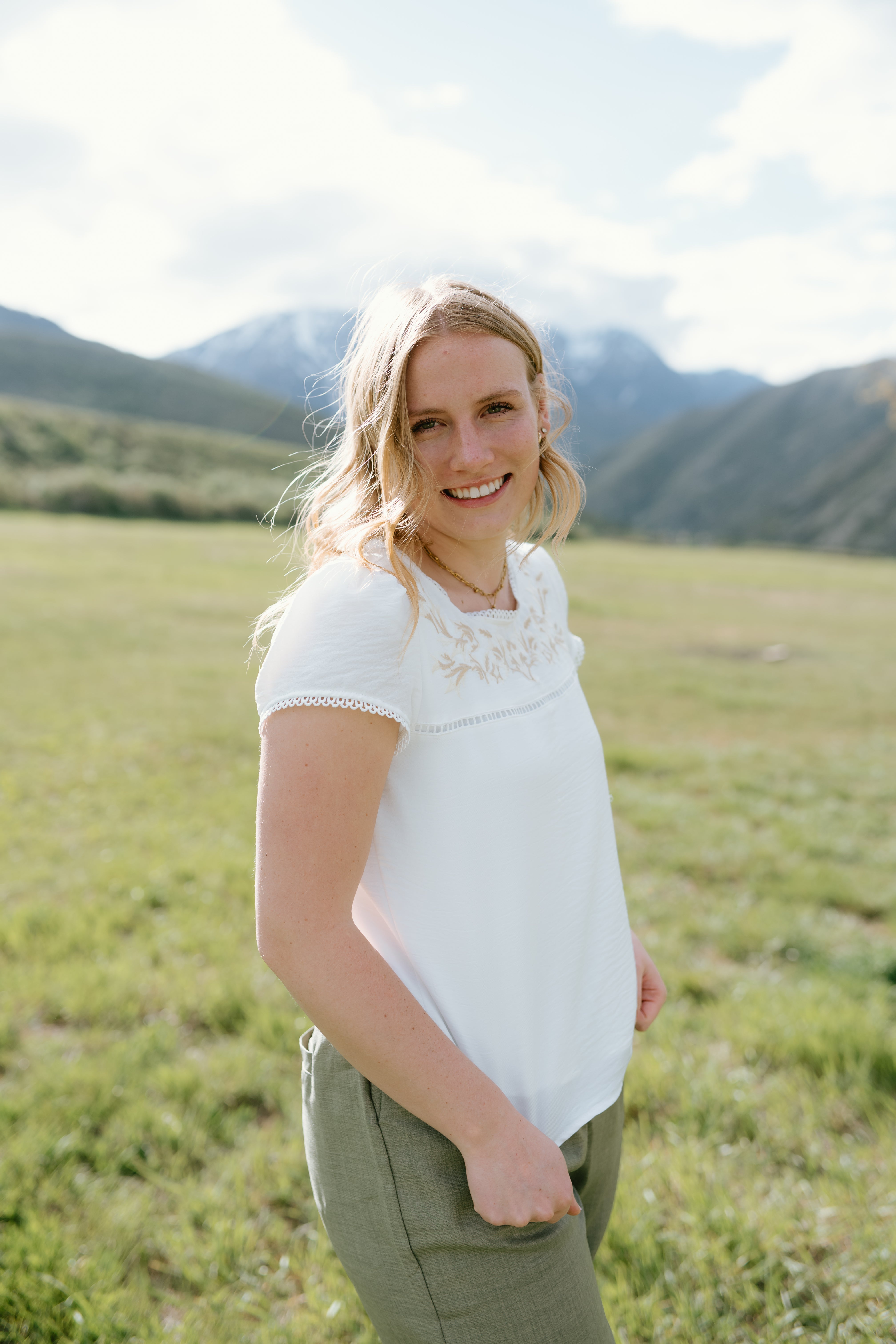 Smiling woman wearing a white Hampton Top with embroidery, standing outdoors in a scenic mountain landscape.