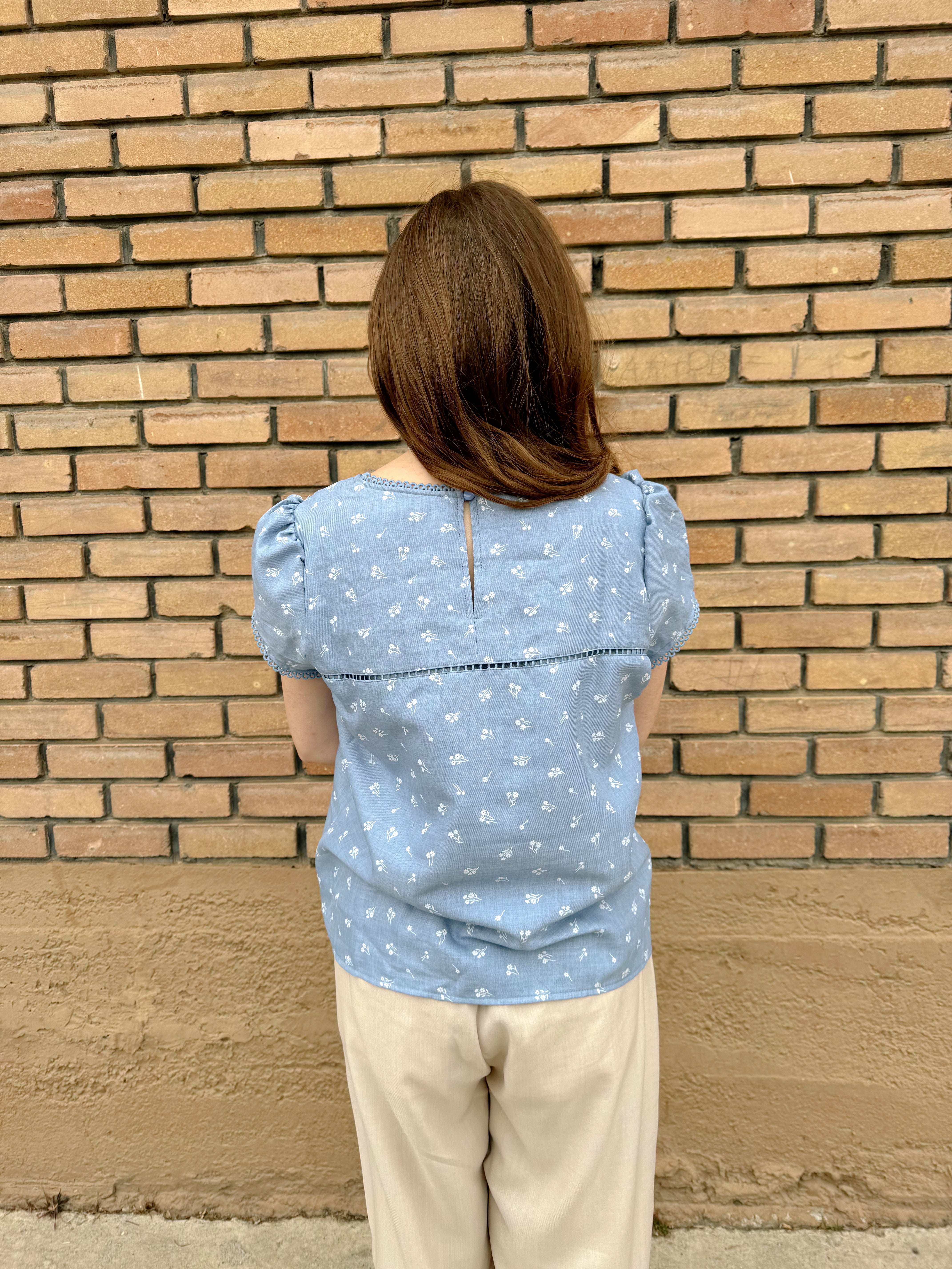 Soft blue Hampton Top with small white floral print, puff sleeves, and lace trim, showcased by a woman standing against a brick wall.