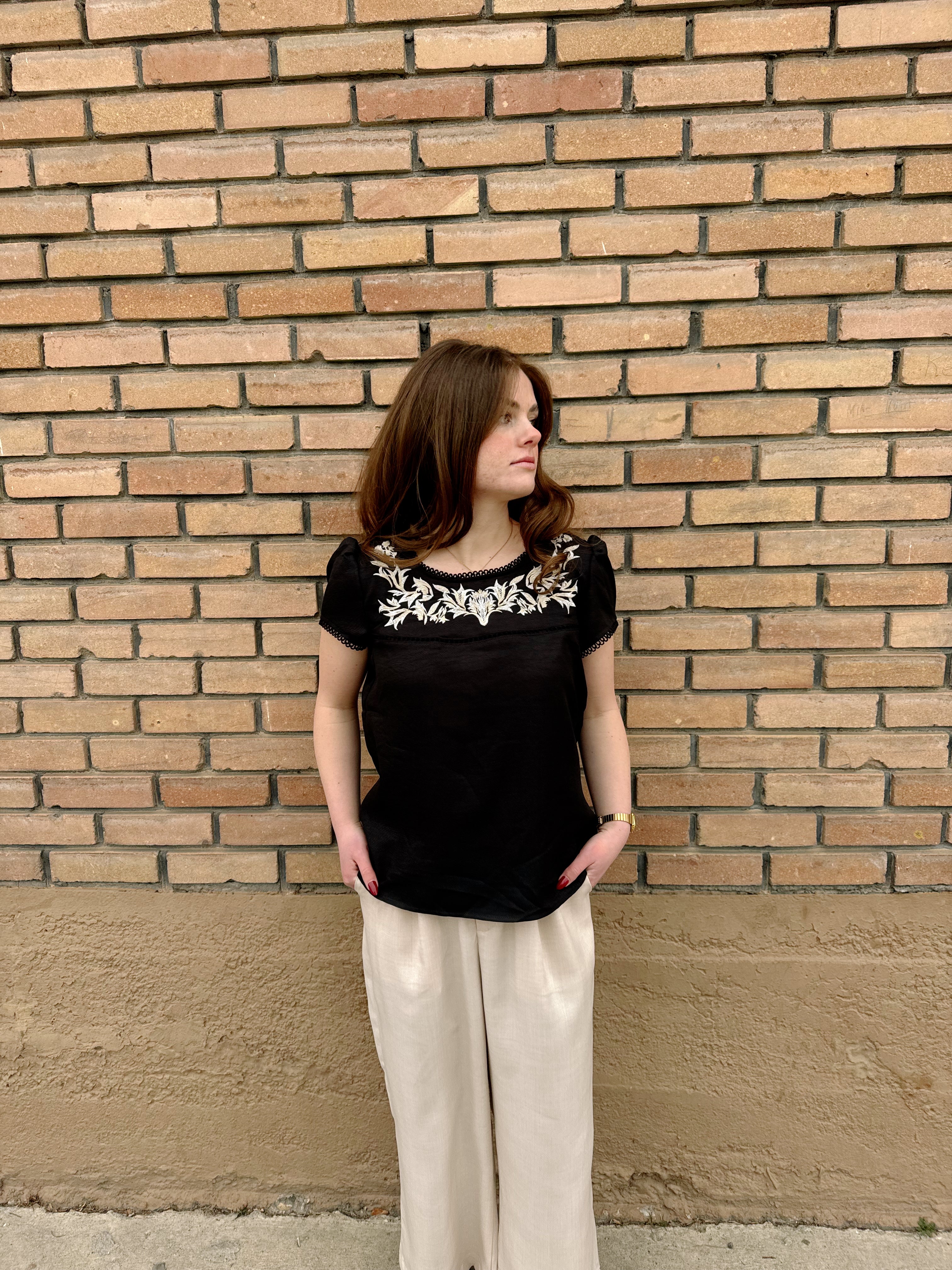 Black Hampton Top with embroidered floral details, worn by a woman against a brick wall.